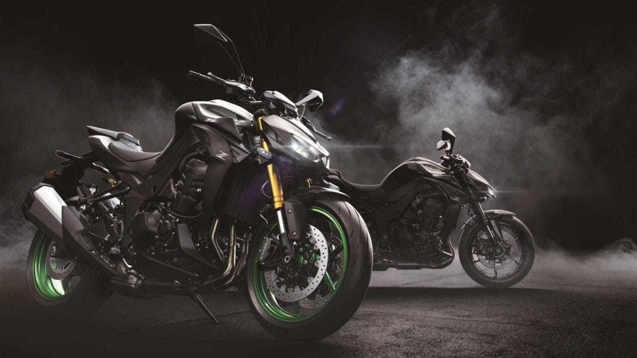 Two sport motorcycles on a dark, smoky backdrop; foreground bike has bright green rims and gold front forks, logo visible on the tank.”] ,