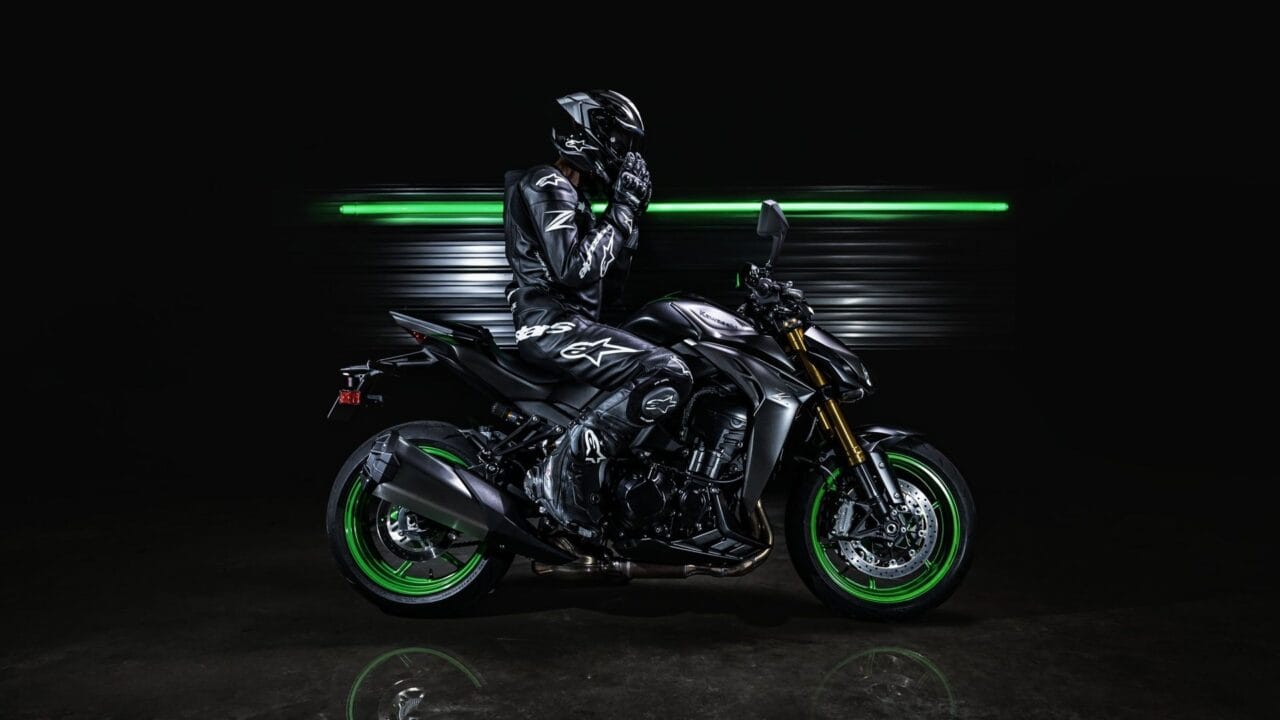 Motorcyclist in black protective gear and helmet sits on a dark sport bike with bright green rims against a dark backdrop.
