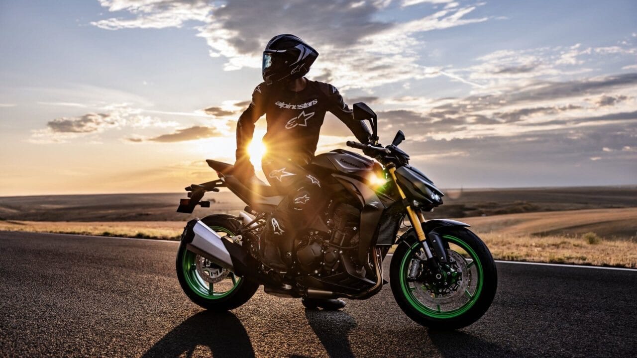 Motorcyclist in full gear sits on a black Kawasaki motorcycle with green rims at sunset on a deserted highway.