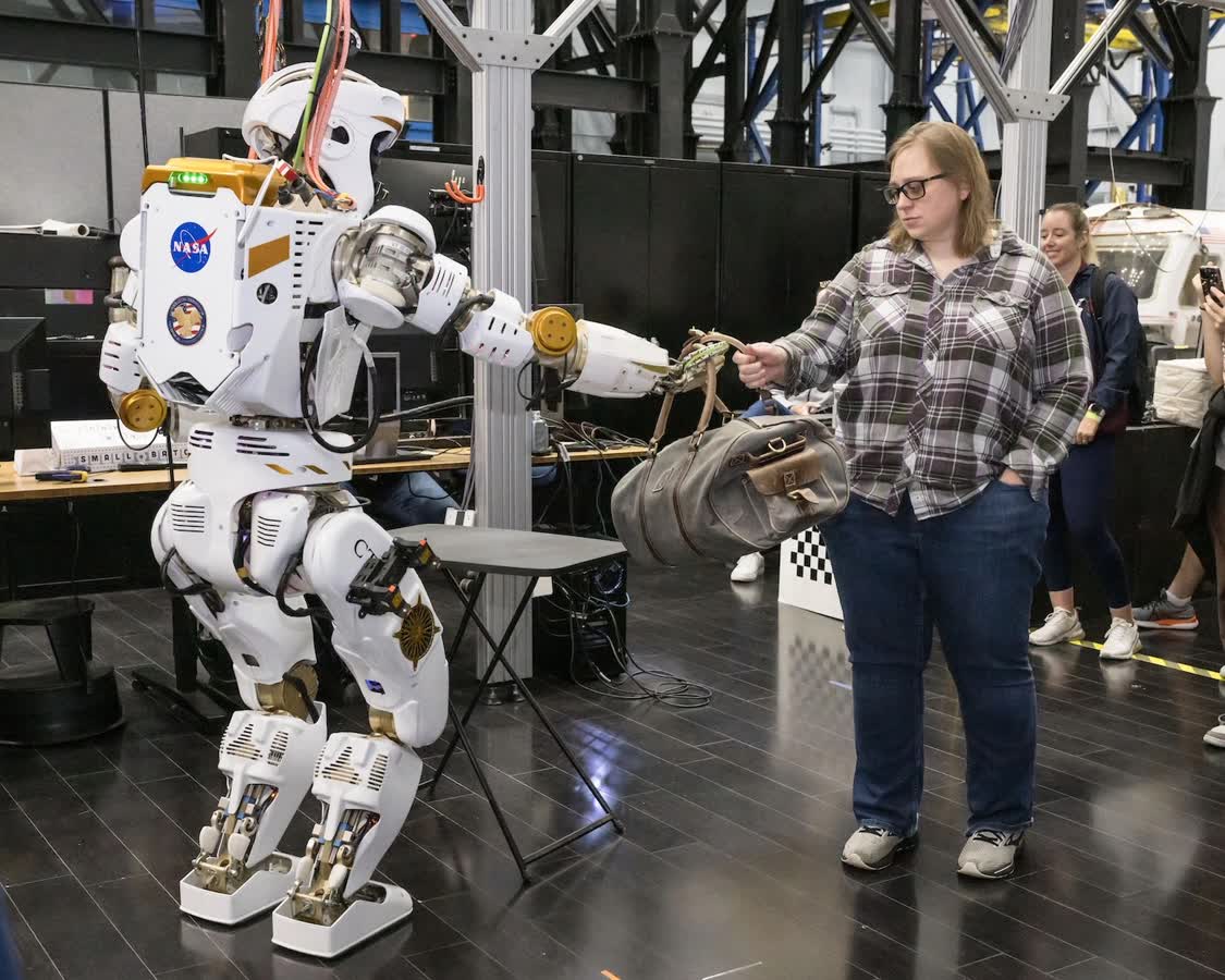 NASA humanoid robot shaking hands with a woman in a lab, cables and equipment visible in the background.