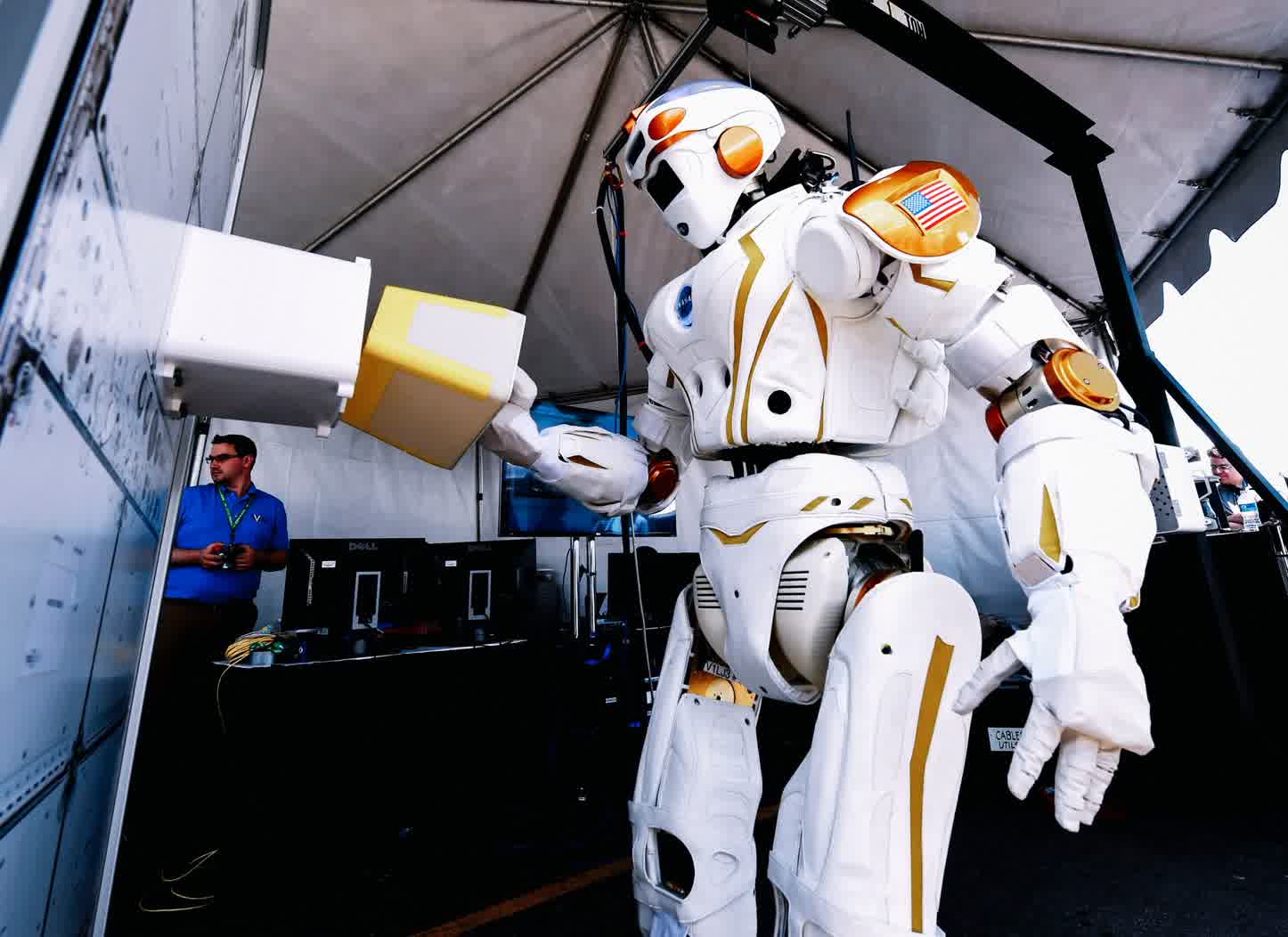 Large white and orange humanoid robot hands a yellow box to a monitor station inside a tent, with a technician watching nearby.
