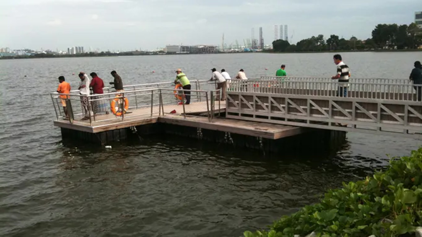 People fishing from a wooden pier that extends into the water, with a life preserver on the railing and an industrial harbor skyline in the distance.