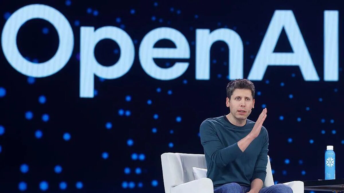 Man in a teal shirt sits on a white chair on stage with a large OpenAI sign behind him; a blue bottle sits on a table beside him.