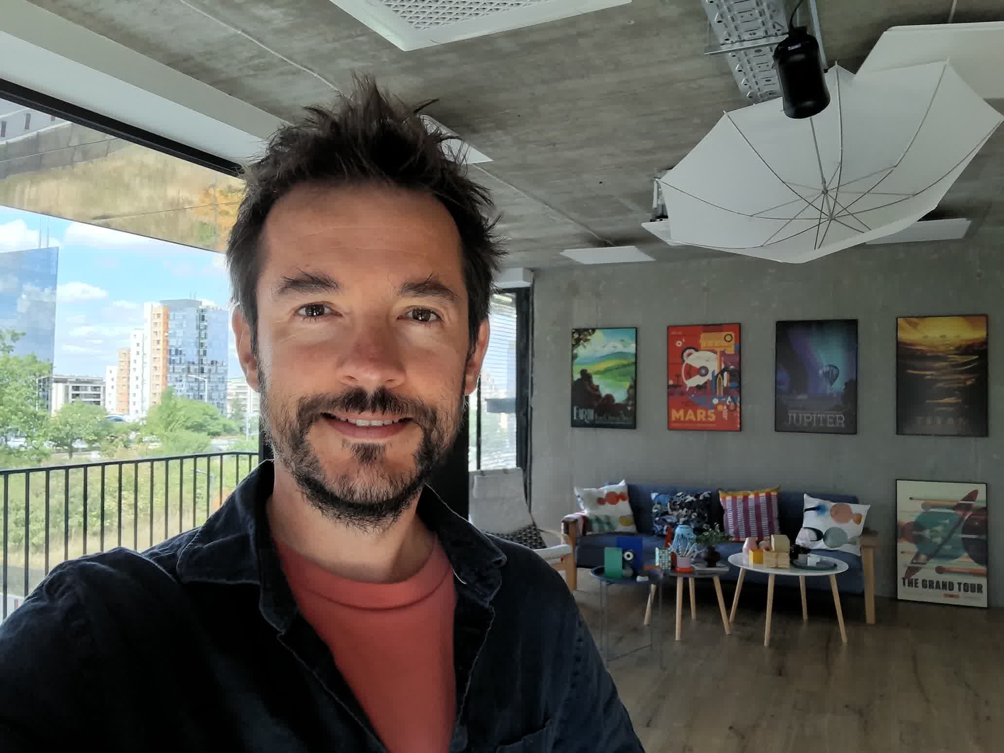 Selfie of a smiling man with short dark hair and stubble indoors, with colorful posters on the wall behind him and a window on the left.
