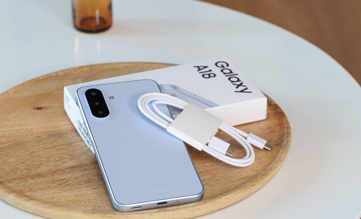 Blue Samsung Galaxy A18 smartphone with a white charging cable and adapter resting on its box on a round wooden tray.