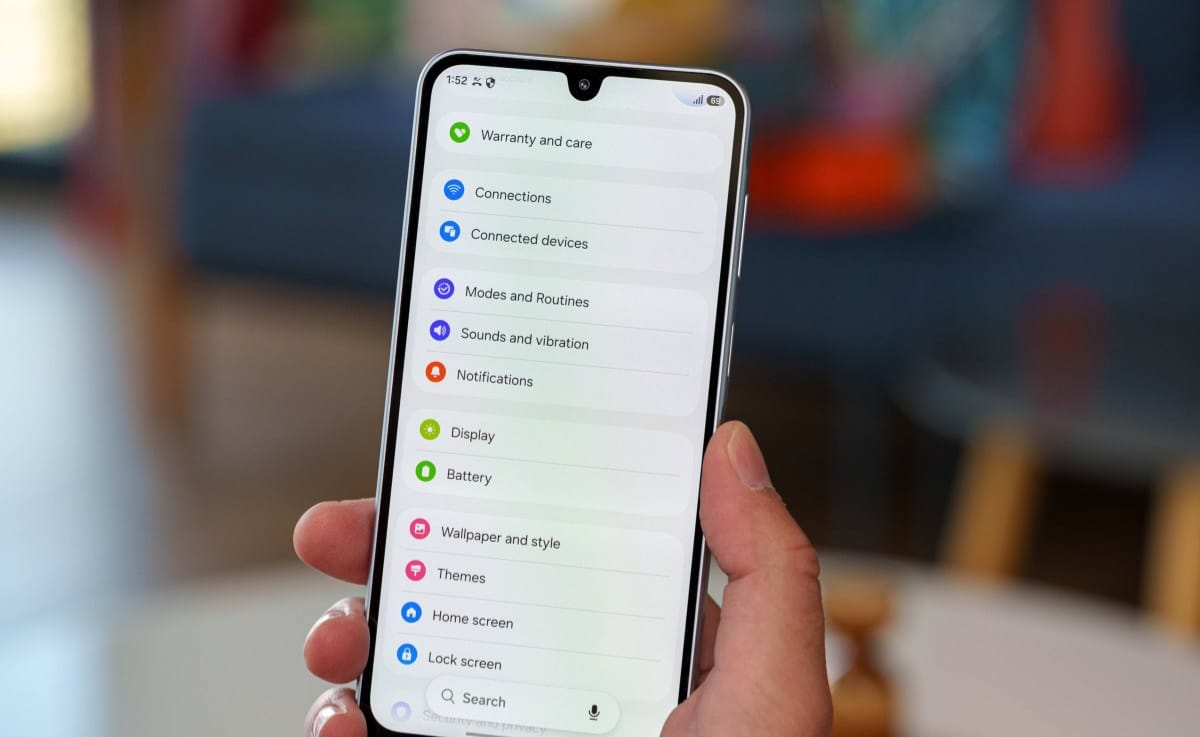 Hand holding a smartphone showing the device’s settings menu (Connections, Display, Battery, Themes, Home screen).
