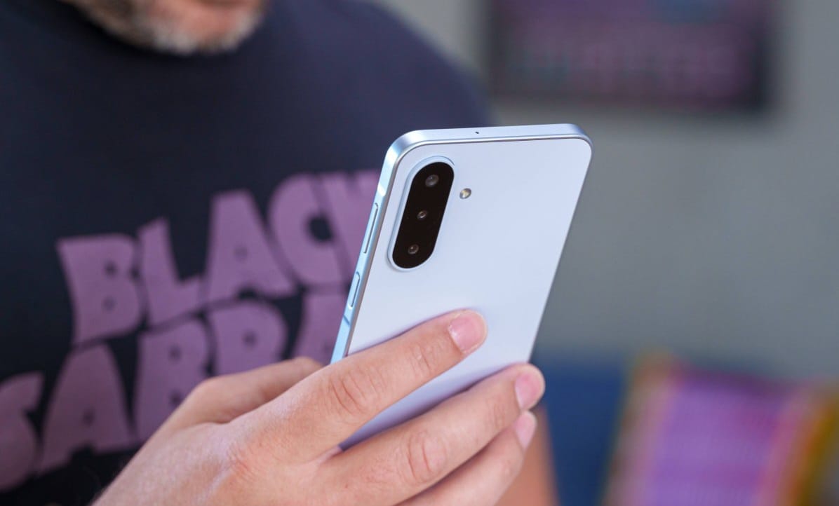 Person in a dark shirt holds a light blue smartphone, rear triple-camera setup visible.
