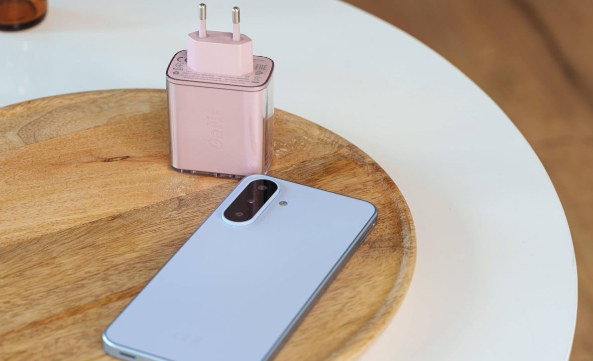 Light blue smartphone with a large camera module rests on a wooden cutting board beside a pink plug-in power adapter.