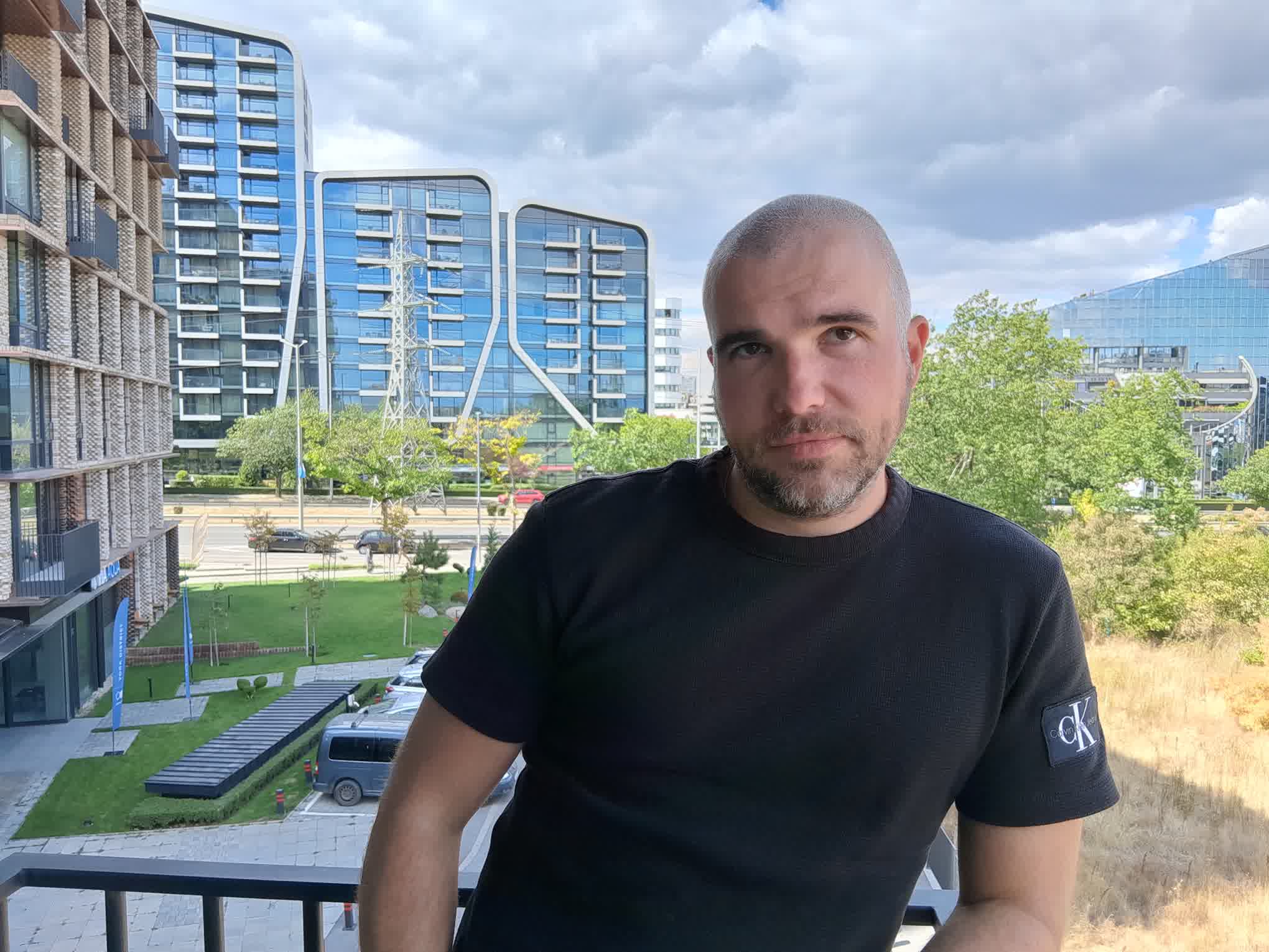 Man in a black t-shirt taking a selfie on a balcony, with blue glass apartment/office buildings behind him under a cloudy sky.