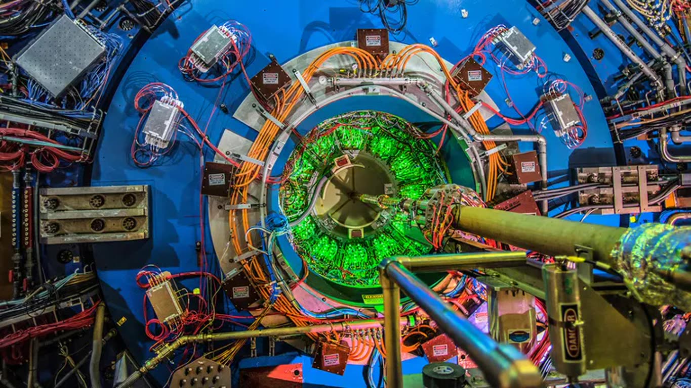 Circular tokamak-like chamber with green-glowing coils, orange cables, and a blue metal frame surrounding the apparatus.