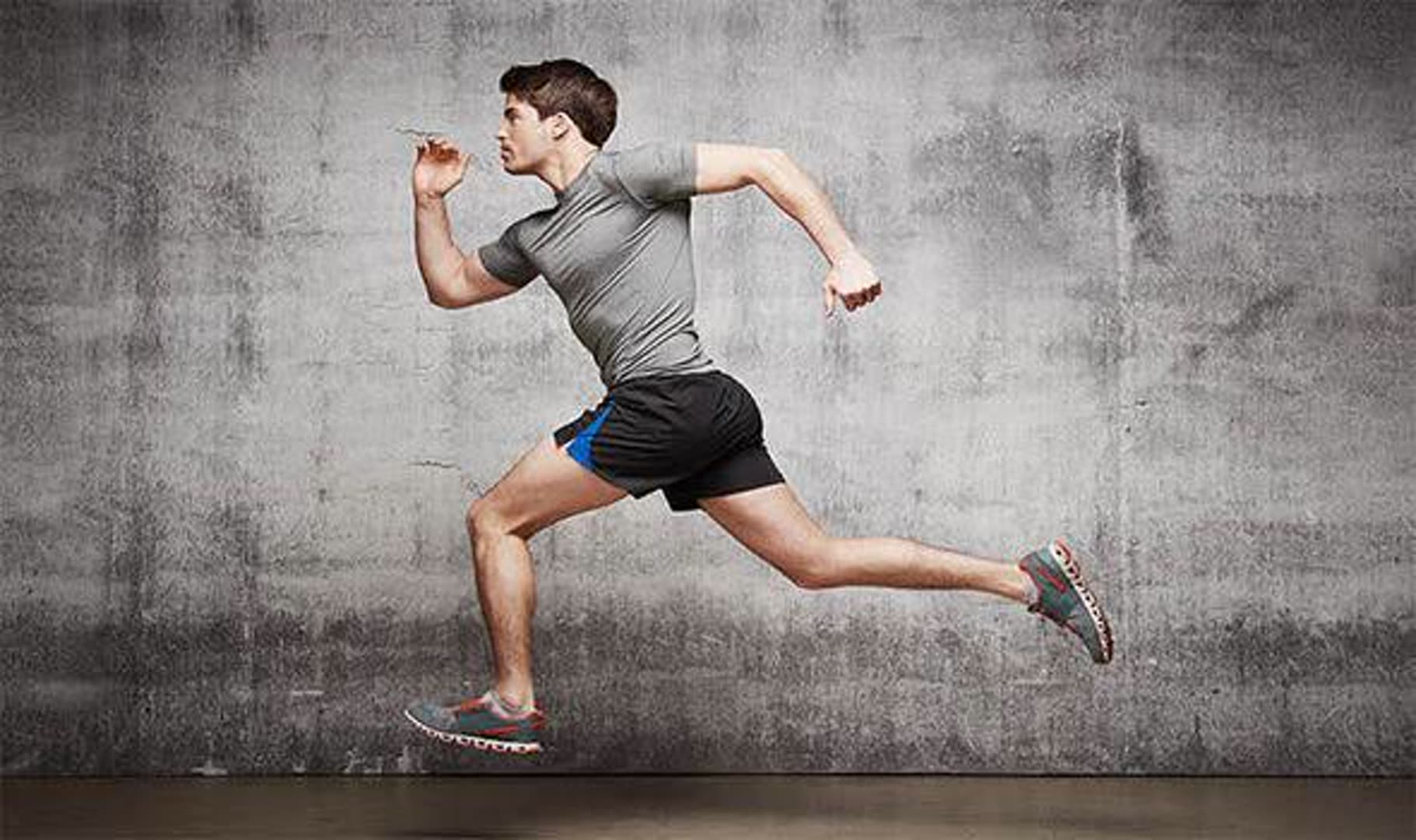 Male runner in a gray athletic shirt and black shorts performs a mid-air sprint leap against a textured concrete wall? (image shows him mid-stride)