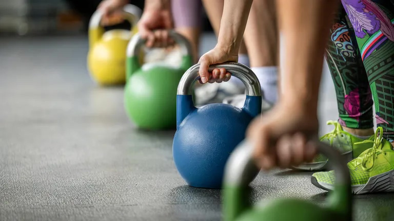 People in a gym performing kettlebell exercises, with a blue kettlebell in the foreground and others in the line behind it.