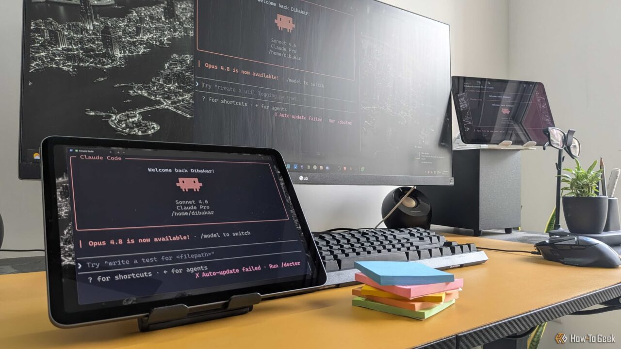 Desk workspace with a tablet and a large monitor showing red-on-black terminal text, plus a keyboard, mouse, and colorful sticky notes.