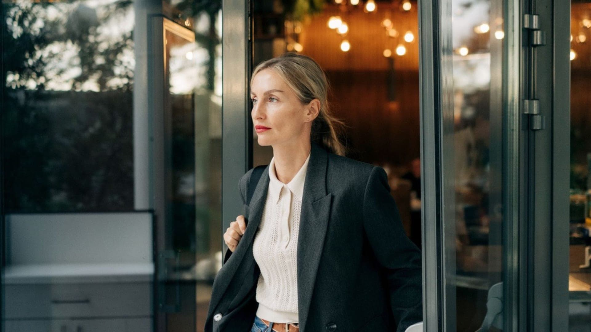 Professional woman in a blazer stepping out through a glass door, carrying a bag over her shoulder.