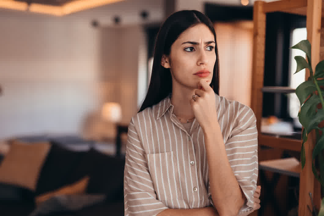 Woman with hand on chin, looking pensive in a cozy indoor setting wearing a striped button-down shirt.