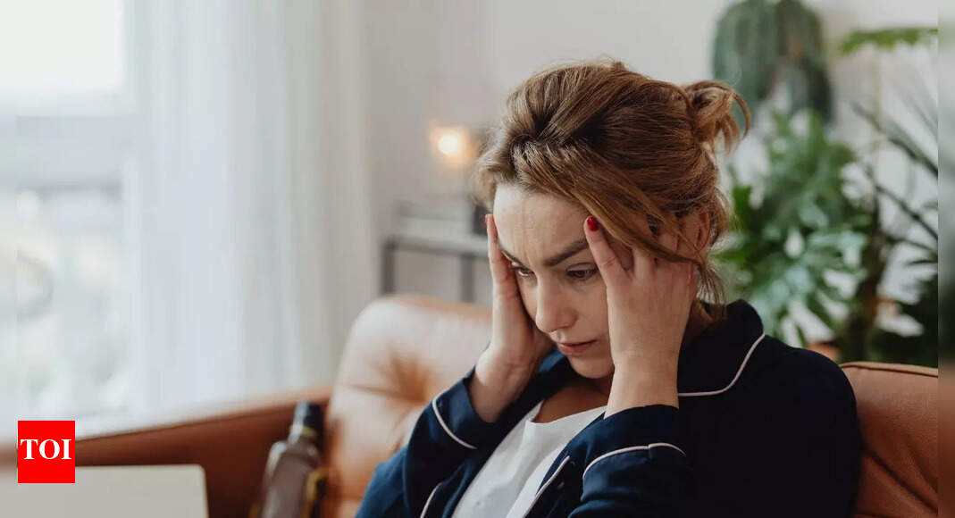 Anxious woman sits on a sofa at home, hands pressed to her temples and a concerned expression on her face.