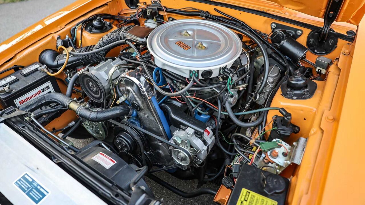 Close-up of an orange car's engine bay with a chrome circular air cleaner, belts, hoses, and a Motorcraft battery.