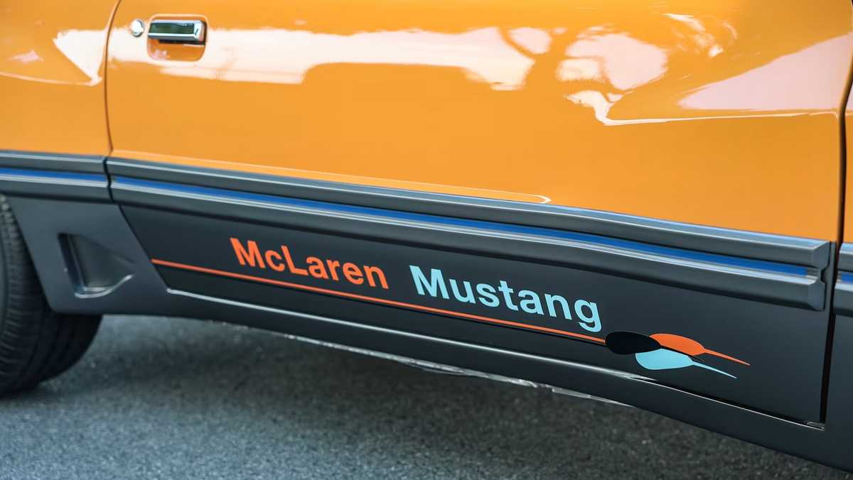 Side view of an orange car with a black lower panel reading ‘McLaren Mustang’ and a teal/orange swoosh.