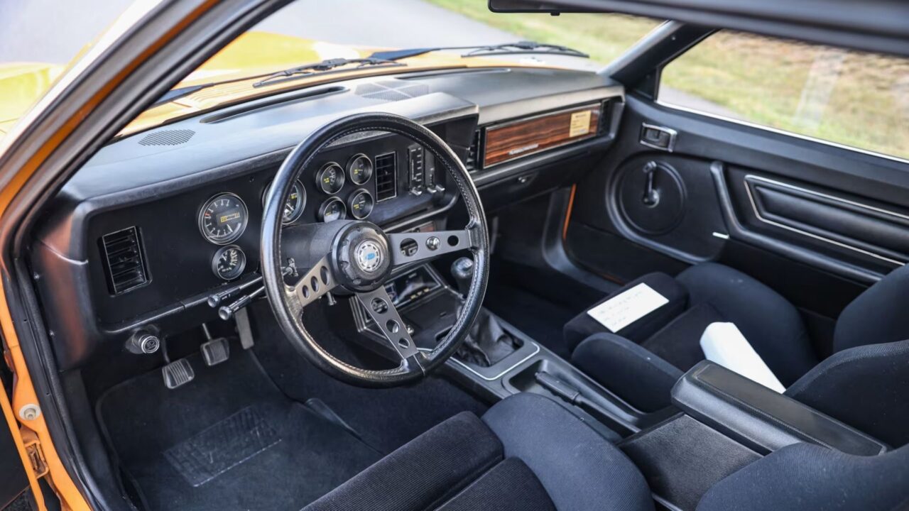 Interior of a vintage car: black dashboard with multiple round gauges, a perforated metal three-spoke steering wheel, and wood trim on the passenger side; orange exterior visible through the door frame.