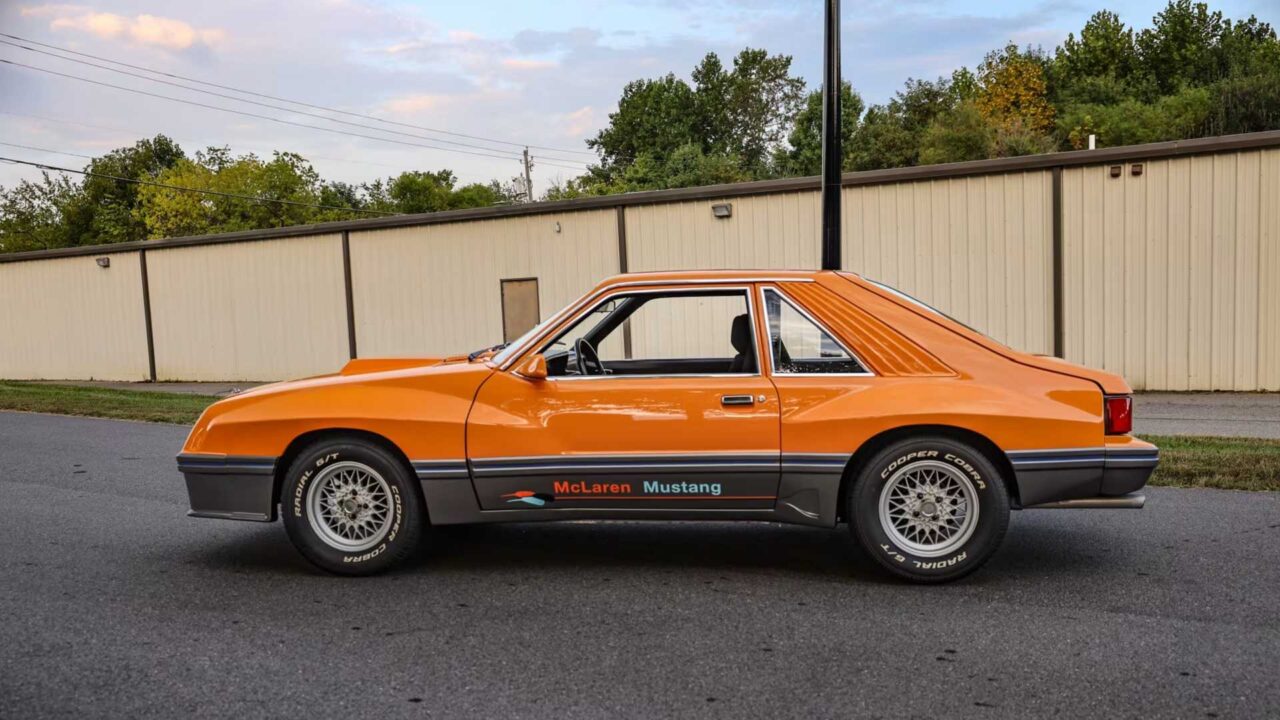 Orange vintage two-door car with racing decals reading 'McLaren Mustang' parked on asphalt near a beige warehouse backdrop.