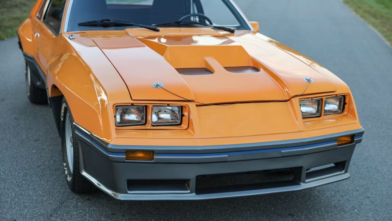 Front view of an orange vintage muscle car, showing its hood scoop, quad headlights, and chrome bumper edge on a road surface, angled from the left front.