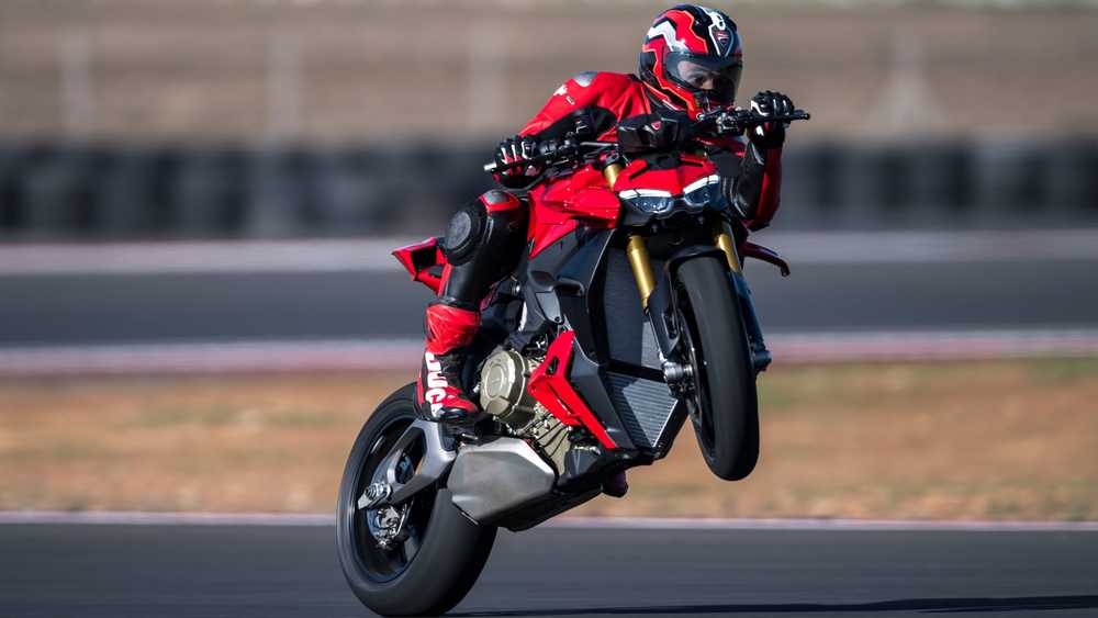 Moto rider in red gear performing a wheelie on a red sport bike on a race track, with the front wheel lifted off the ground.