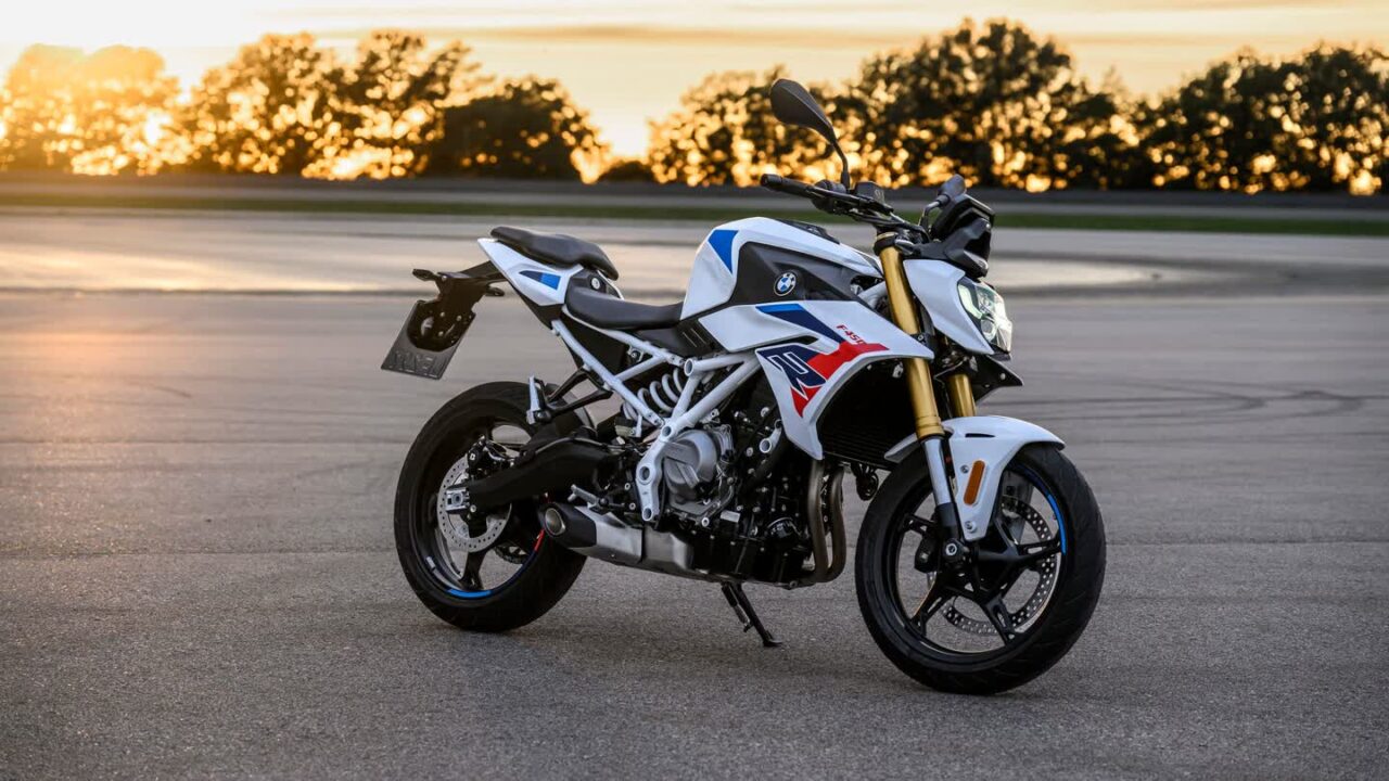 White BMW sport motorcycle with blue and red graphics on a road at sunset, resting on its kickstand with trees glowing in the background.