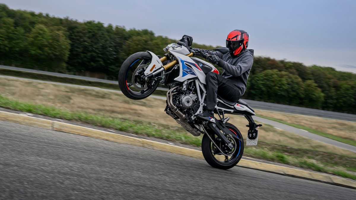 Rider in a red helmet performing a wheelie on a white/blue sport motorcycle on a paved road.