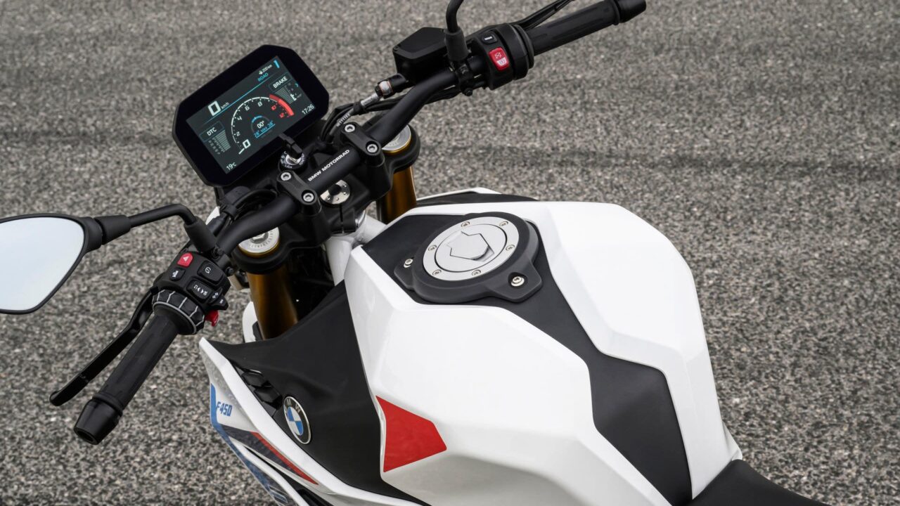 Close-up of a white BMW motorcycle tank and handlebars with a digital dashboard display visible to the left of the fork blend.