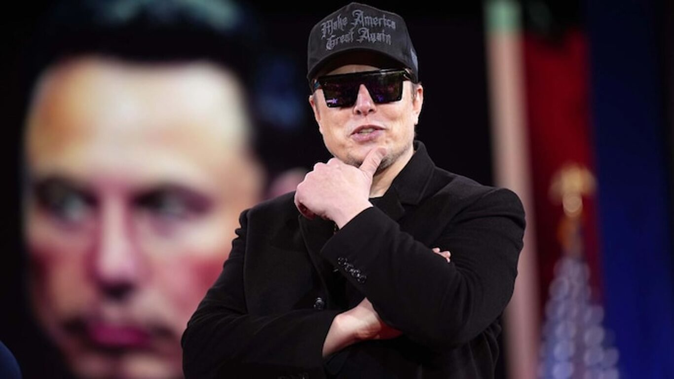 Man in a black blazer and sunglasses, wearing a MAGA cap, poses with hand on chin in thoughtful gesture against a dark background.