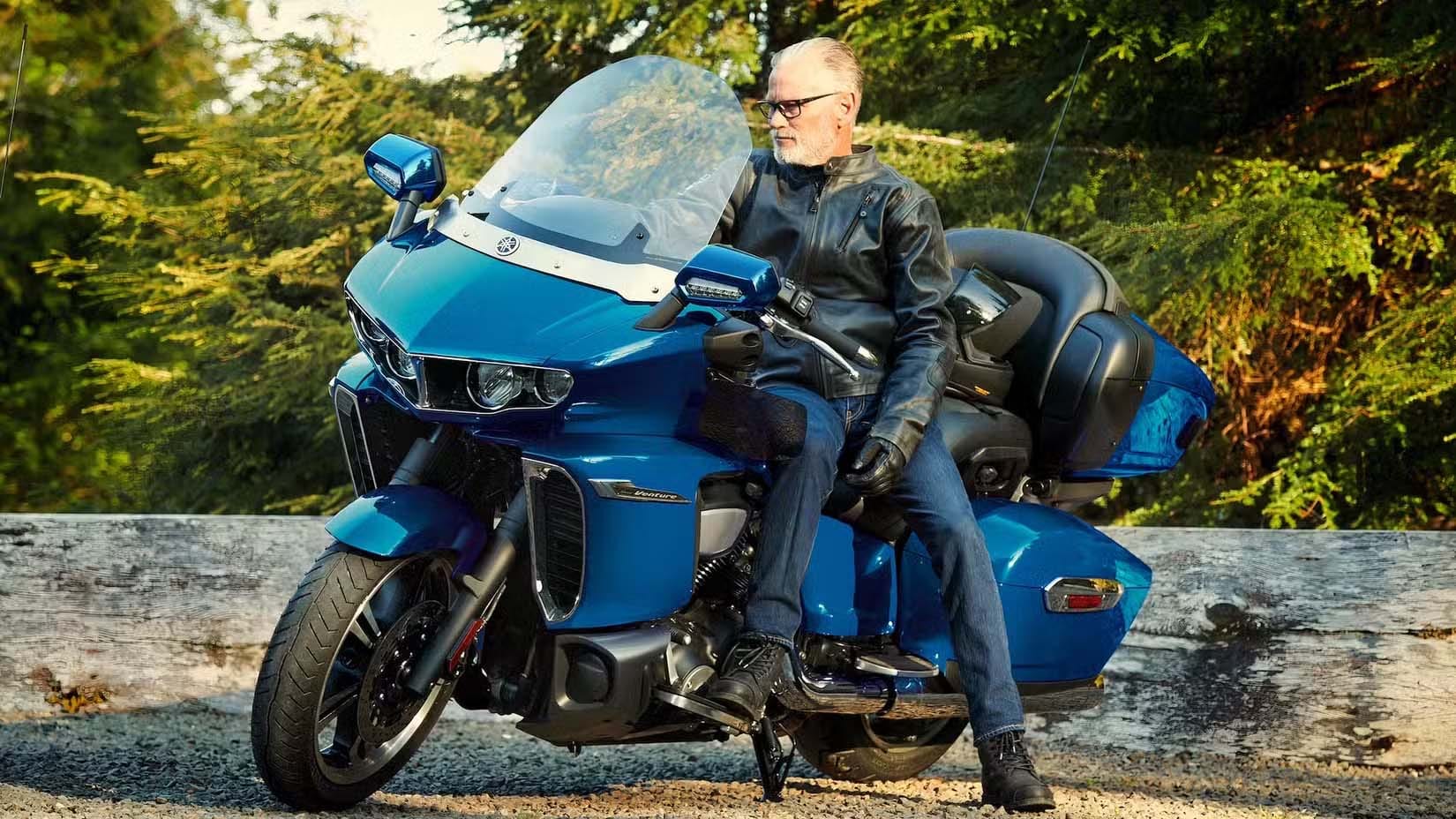 Man in a leather jacket sits on a large blue touring motorcycle with a windshield and storage, beside a wooden barrier.