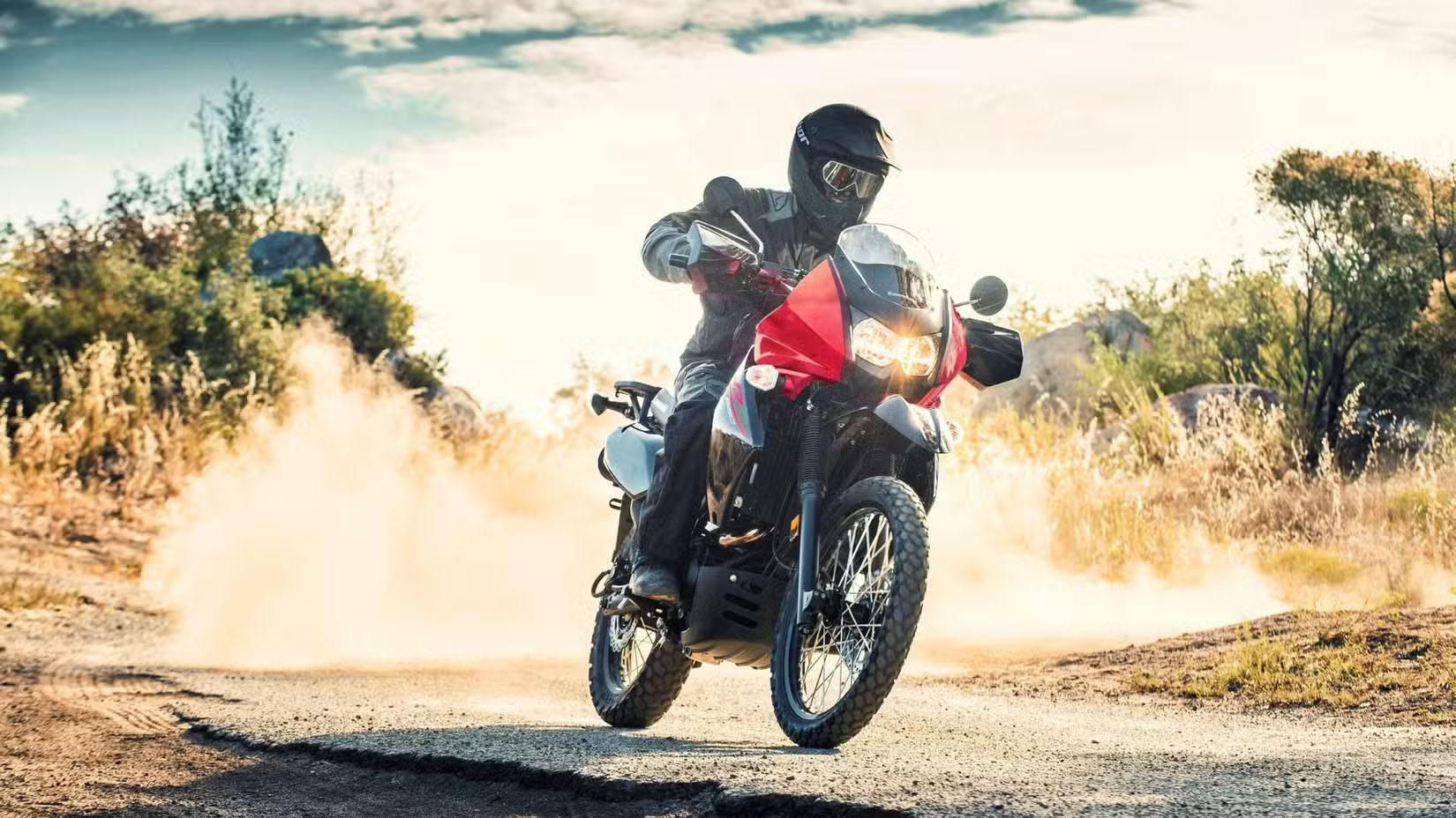 Motorcyclist in protective gear riding a red dual-sport motorcycle on a dusty dirt road, kicking up a cloud of dust behind them in a dry landscape with shrubs and rocks nearby.