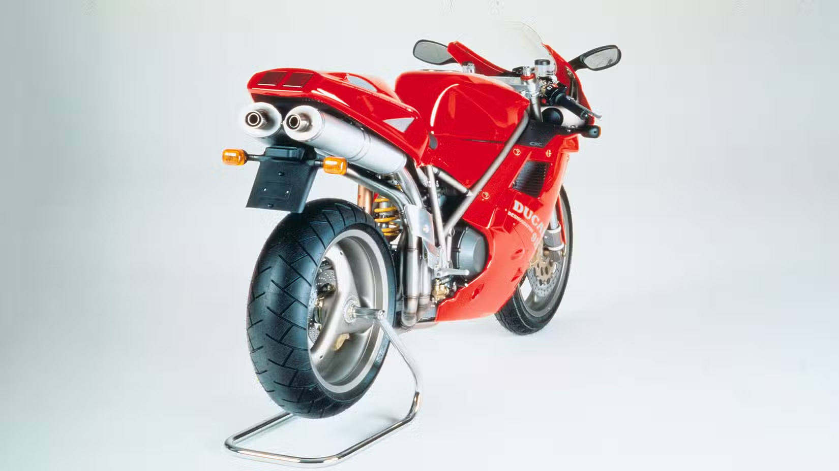 Red Ducati sport motorcycle on a metal stand in a light studio background, angled rear view.