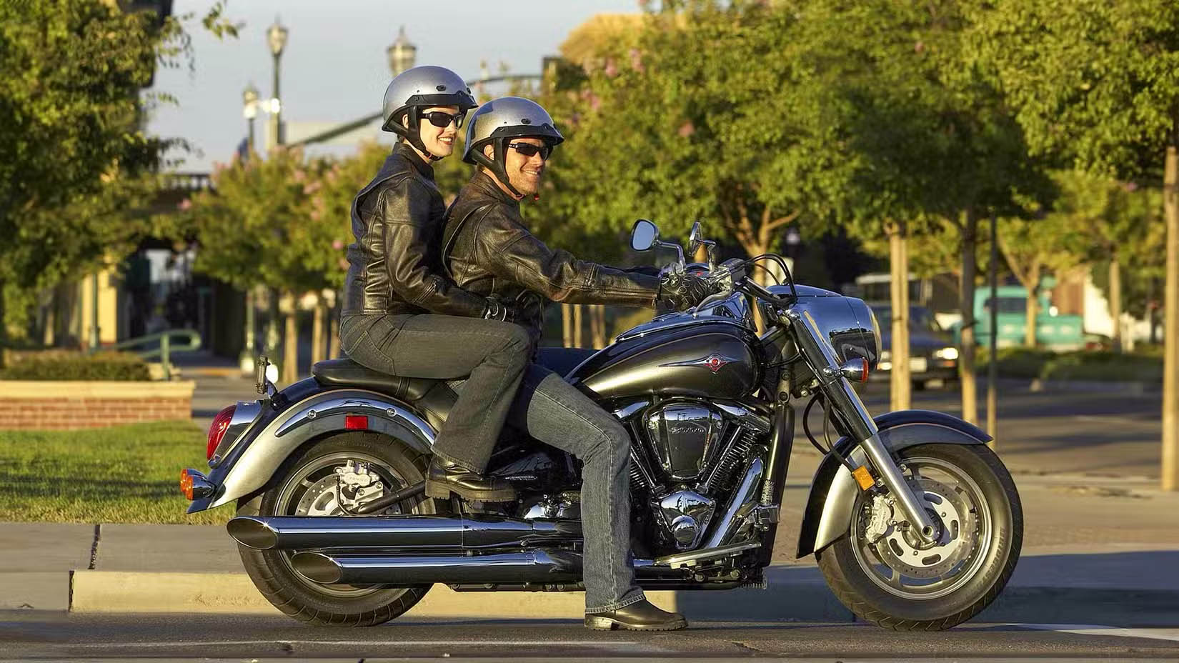 Couple riding a black touring motorcycle in leather jackets and helmets on a sunny street.