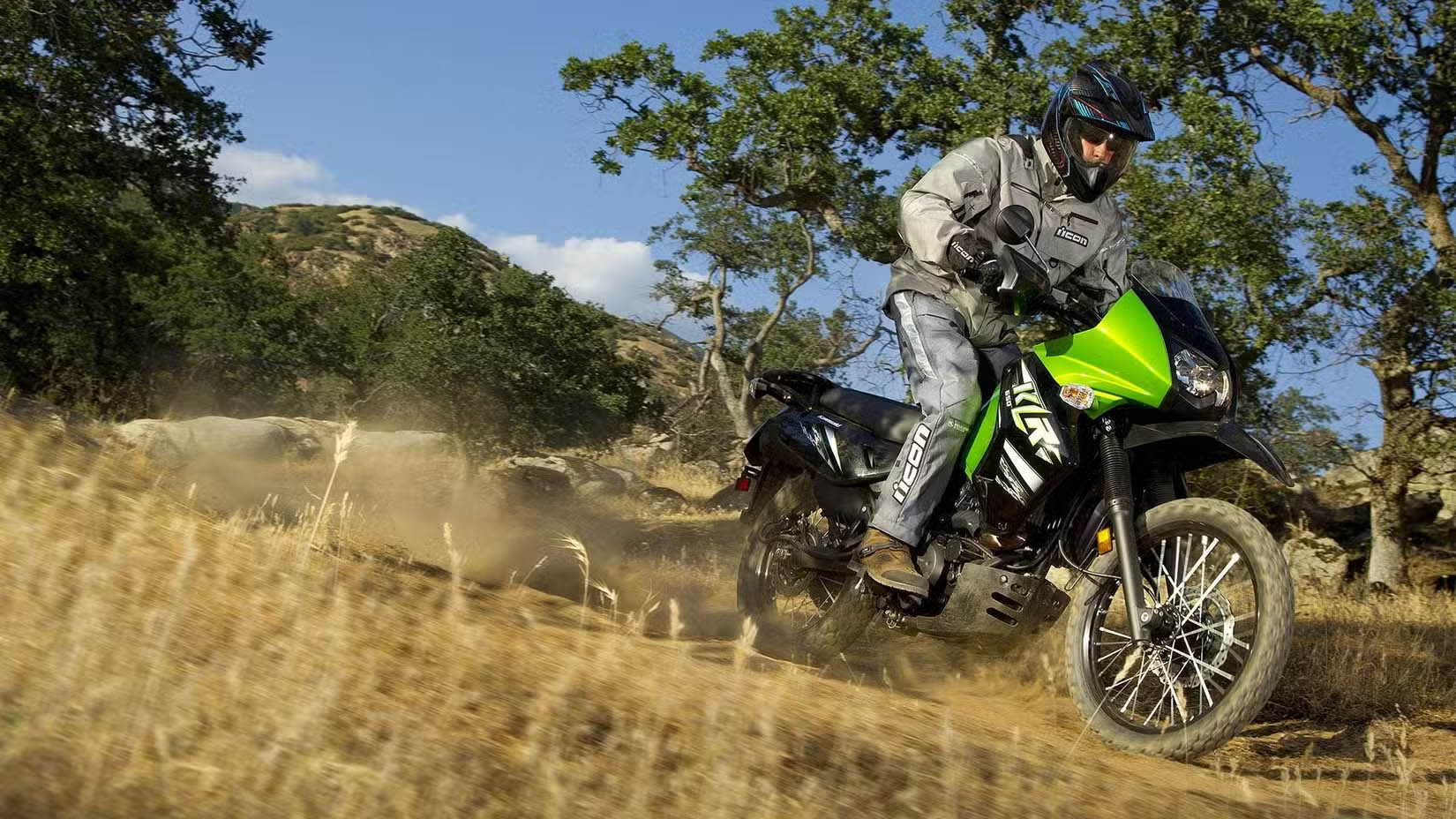 Motorcycle rider in protective gear navigating a dusty dirt trail through a hillside landscape.