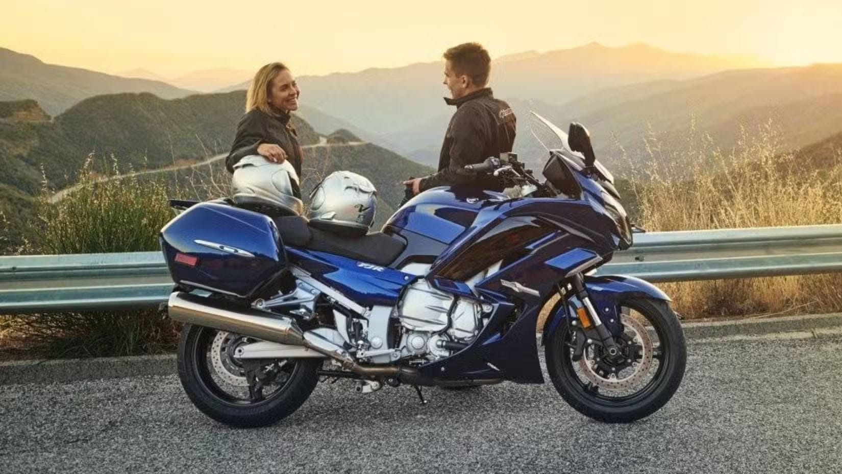 Two motorcyclists stand beside a blue sport motorcycle on a mountain road at sunset, wearing jackets with two helmets resting on the seat.