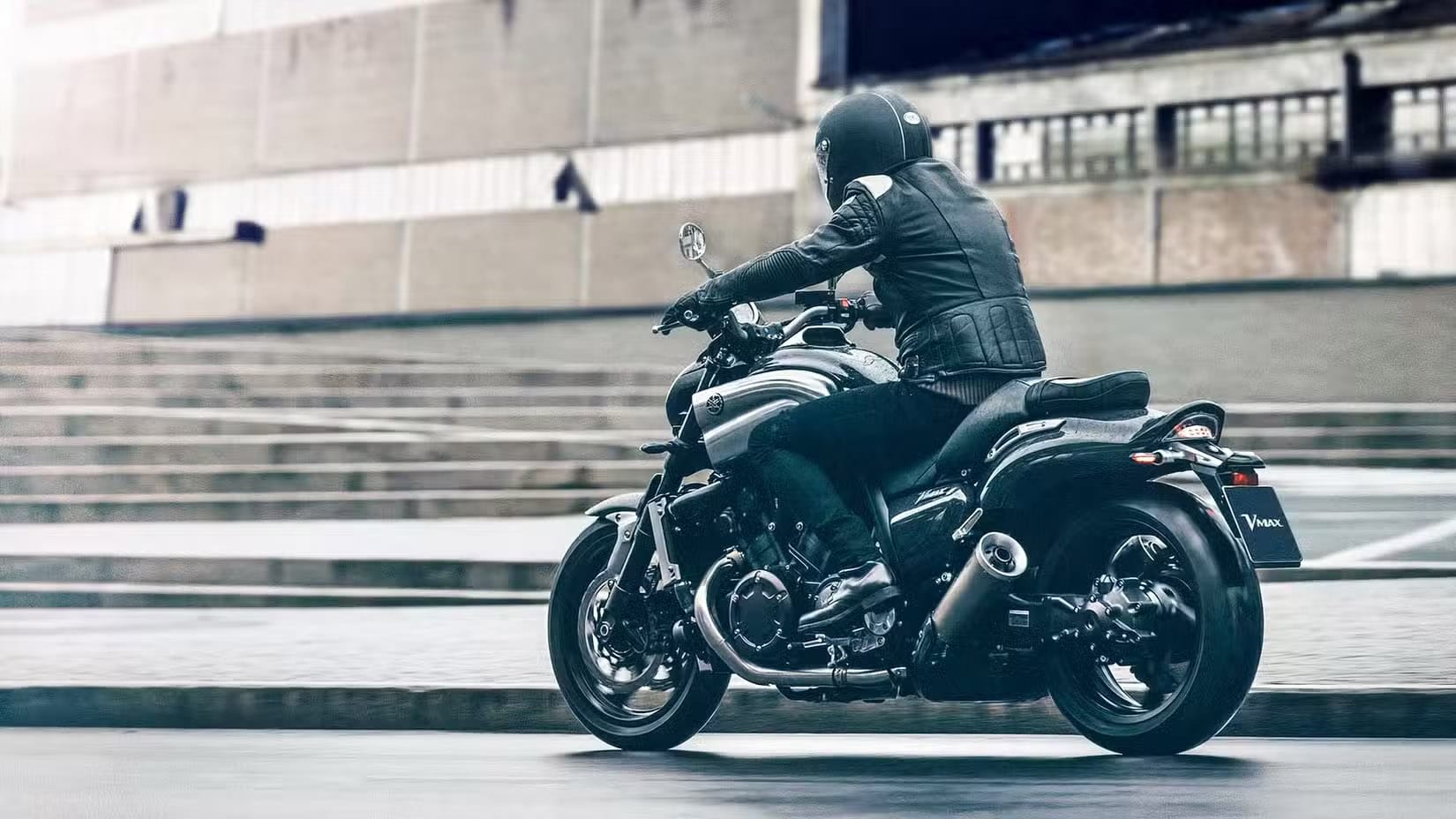 Motorcyclist in black gear riding a Yamaha VMAX motorcycle on a city street with concrete barriers in the background