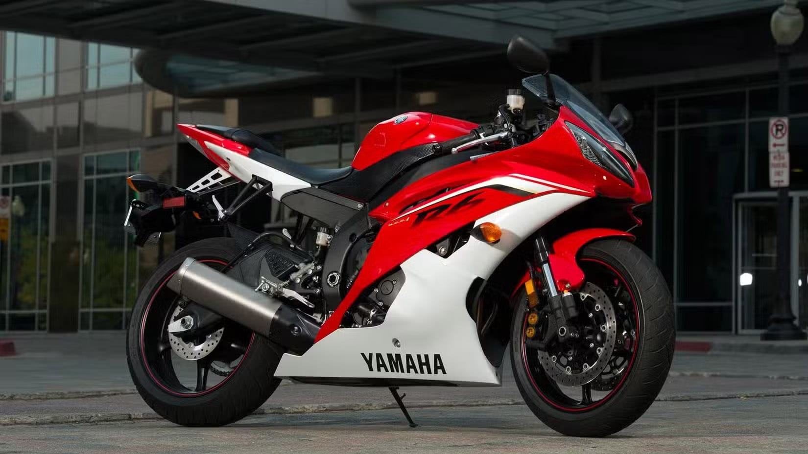 Red and white Yamaha sport motorcycle parked on a city street with a modern glass building in the background.