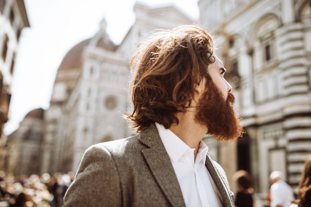 Side profile of a bearded man with wavy brown hair wearing a gray blazer on a sunny city street with historic buildings in the background.