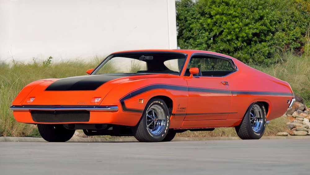 Orange vintage muscle car with black hood stripe parked on a street; side stripe along the doors and chrome wheels visible.