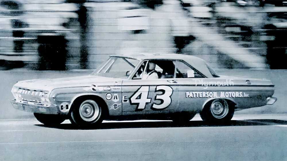 Black-and-white photo of a vintage race car, number 43, speeding on a track with 'PATTERSON MOTORS' lettering along the side.