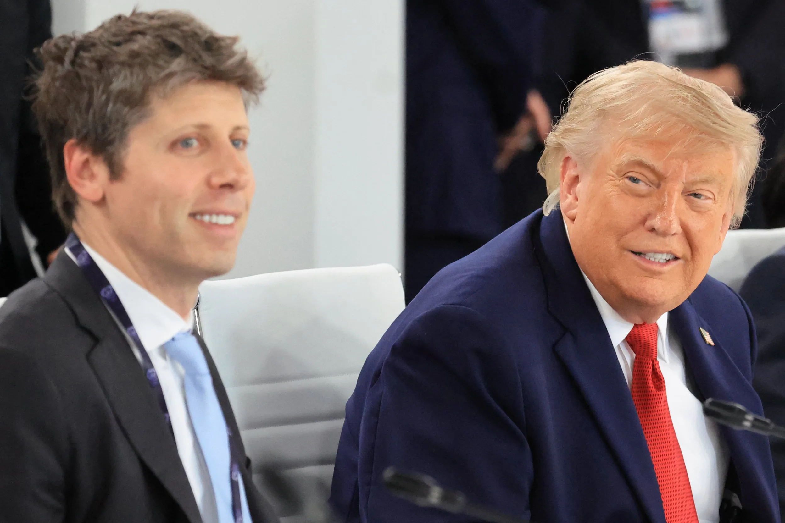 Two men in suits sitting indoors; on the right a man in a navy suit with a red tie smiles at the camera, while the left man looks sideways.
