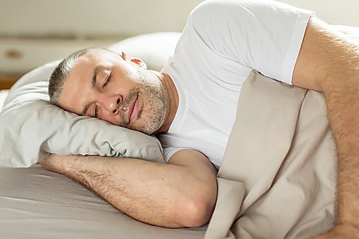 Man sleeping on his side with a pillow under his head in a bed, wearing a white t‑shirt and beige blanket nearby