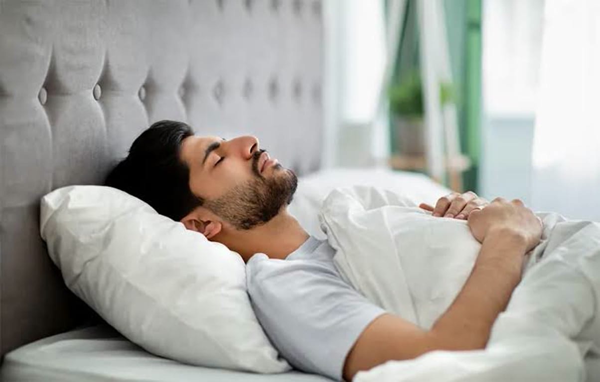 Man sleeping peacefully in bed with white sheets, resting against a tufted headboard in a bright bedroom