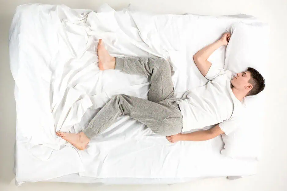 Top-down view of a man lying on a white bed wearing a white T-shirt and gray joggers.