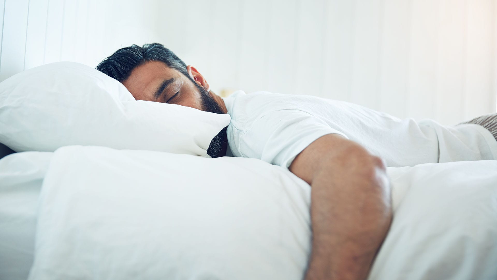 Bearded man sleeping on a white pillow in a bright bedroom.