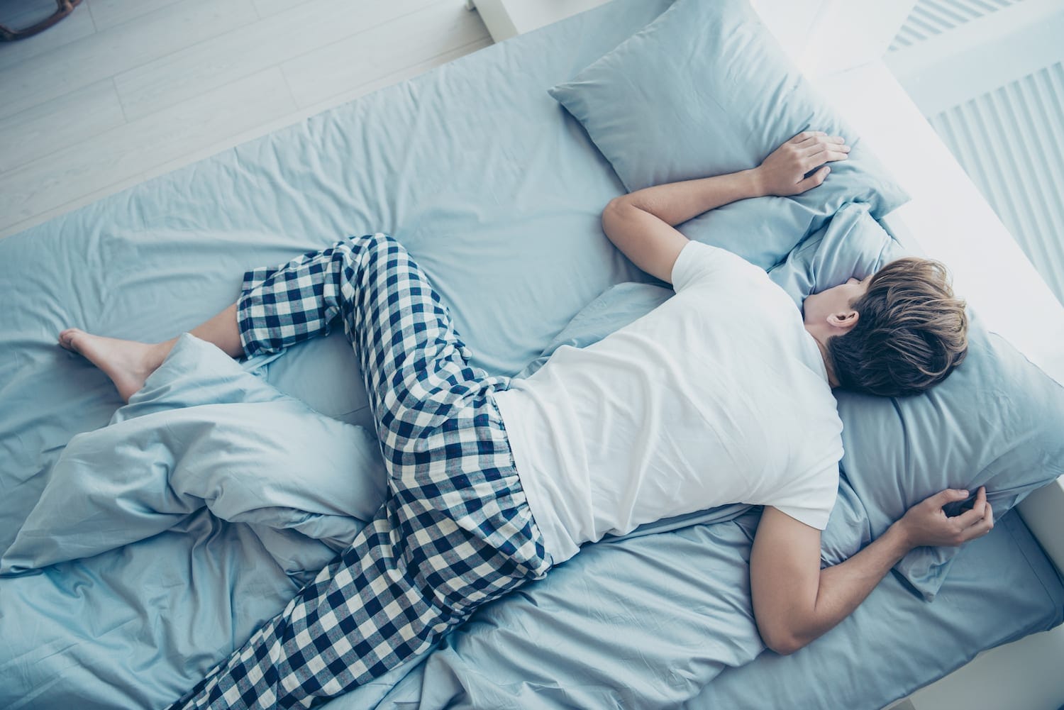 Person lying face down on a light blue bed, wearing a white shirt and blue plaid pajama pants, resting on pillows.
