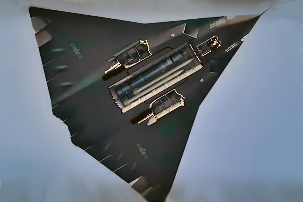 Top-down view of a B-2 Spirit stealth bomber with external ordnance loaded, flying in a clear blue sky.