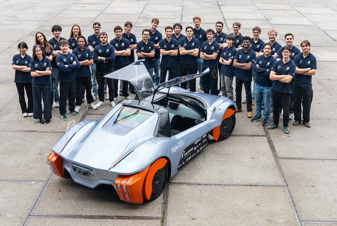 Group of students in navy polo shirts posing beside a silver sports car with orange wheel covers and open gull-wing doors.