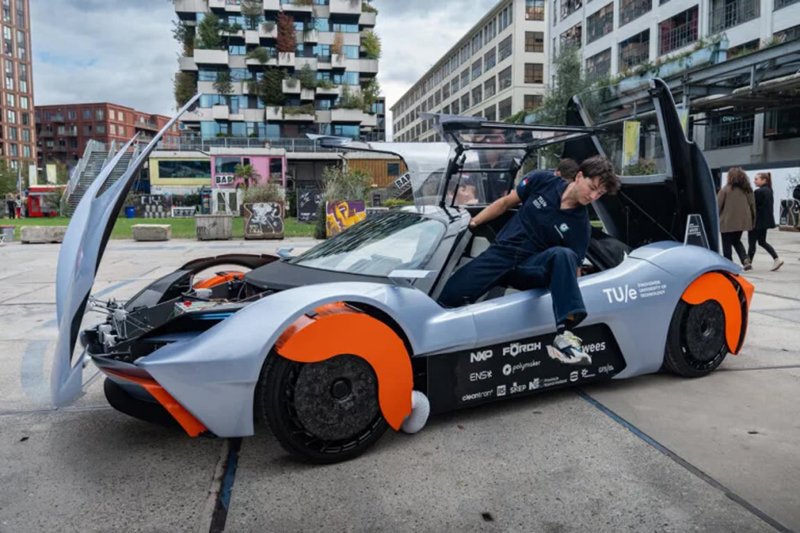 Futuristic silver and orange sports car with doors open, a person in navy overalls stepping into the cockpit in an urban plaza.